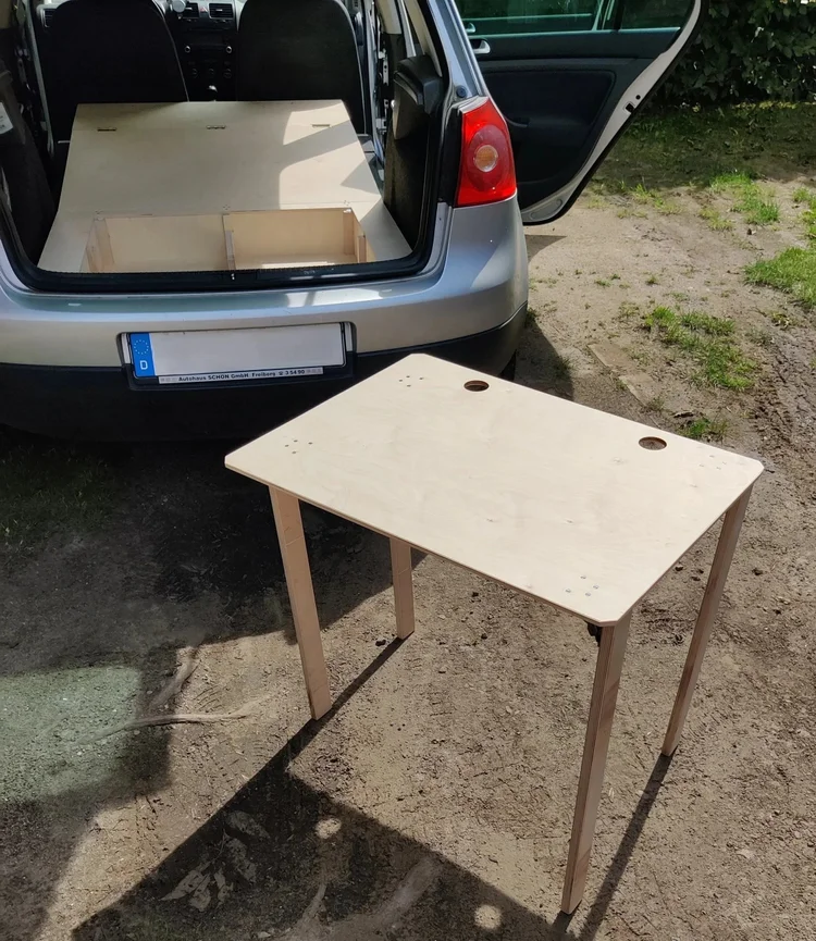 Aufgebauter Campingtisch aus Birkenholz neben dem geöffneten Kofferraum eines VW Golf