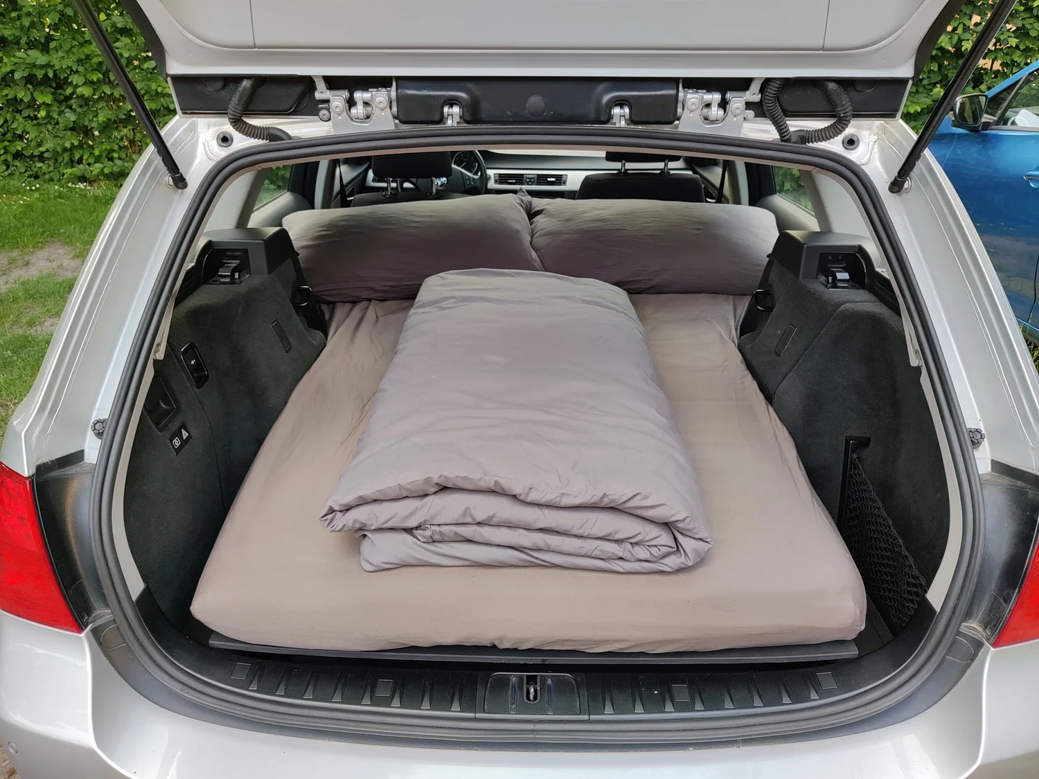 Gemachtes Bett mit Kissen und Decke im Kofferraum eines silbernen BMW Kombi – die Rost Box als Mini-Camper